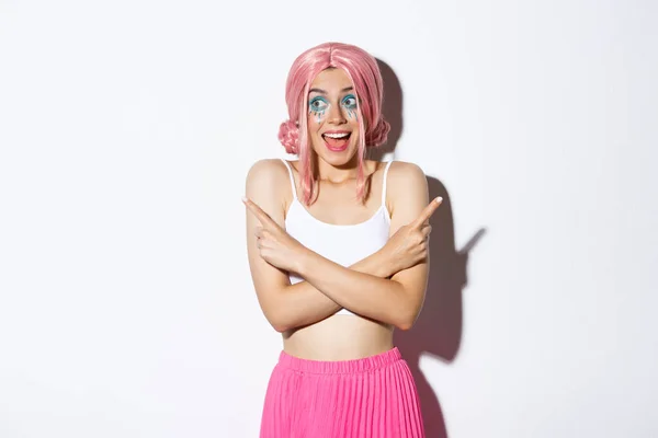 Retrato de menina sorridente animado com peruca rosa e maquiagem festa, apontando os dedos para o lado, mas olhando para a esquerda com expressão feliz, de pé sobre fundo branco — Fotografia de Stock