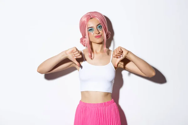 Imagem da jovem cética com peruca rosa e maquiagem brilhante, mostrando polegares para baixo decepcionado, não gosta de comemoração halloween, de pé sobre fundo branco — Fotografia de Stock