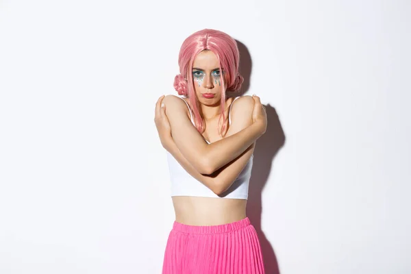 Portrait of offended cute girl hugging herself and sulking, feeling insulted or sad about something, wearing pink wig for halloween celebration, standing over white background — Stok Foto