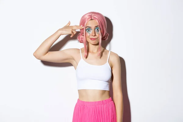 Porträt eines genervten Partygirls mit pinkfarbener Perücke, das Fingergewehrzeichen über dem Kopf macht und irritiert grinst, im Halloween-Kostüm vor weißem Hintergrund stehend — Stockfoto