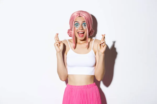 Imagem da menina festa animado cruzando os dedos para a boa sorte e sorrindo espantado, olhando esperançoso, vestindo traje de Halloween com peruca rosa e maquiagem brilhante — Fotografia de Stock