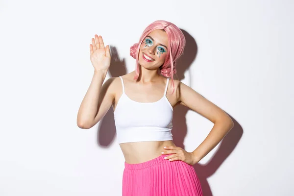 Imagem da menina glamour alegre com peruca rosa e maquiagem halloween, dizendo olá, acenando com a mão e cumprimentando alguém, de pé sobre fundo branco — Fotografia de Stock