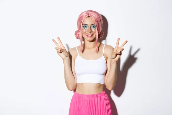 Mulher sorridente bonita celebrando halloween em peruca rosa, mostrando sinais de paz, de pé sobre fundo branco — Fotografia de Stock