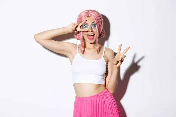Podekscytowany uśmiechnięta ładna dziewczyna w różowej peruce, pokazując znaki pokoju i wyglądając zdumiony, stojąc w kostiumie halloween i różowej peruce — Zdjęcie stockowe