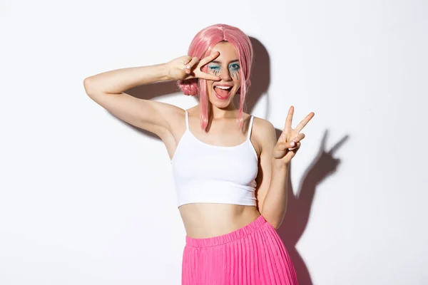 Menina bonita feliz em peruca rosa, vestida-up para festa de Halloween, mostrando sinais de paz, sorrindo e piscando coquete para a câmera — Fotografia de Stock