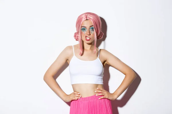 Retrato de mulher atraente atrevida com peruca de anime rosa, maquiagem brilhante, mostrando dentes e olhando confiante, celebrando halloween, de pé sobre fundo branco — Fotografia de Stock
