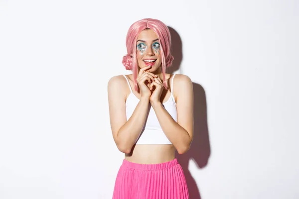 Retrato de linda garota intrigada com peruca rosa e maquiagem brilhante, olhando para a esquerda em algo tentador, de pé em traje de Halloween sobre fundo branco — Fotografia de Stock