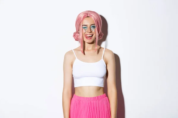 Портрет беззаботной улыбающейся модели в розовом парике вечеринки, подмигивание и показ языка, празднование Хэллоуина, стоя на белом фоне — стоковое фото