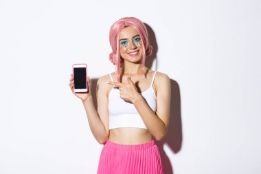 Pembe peruklu ve parlak makyajlı mutlu kadın modelin portresi, cep telefonu ekranını işaret eden, uygulama veya afiş gösteren