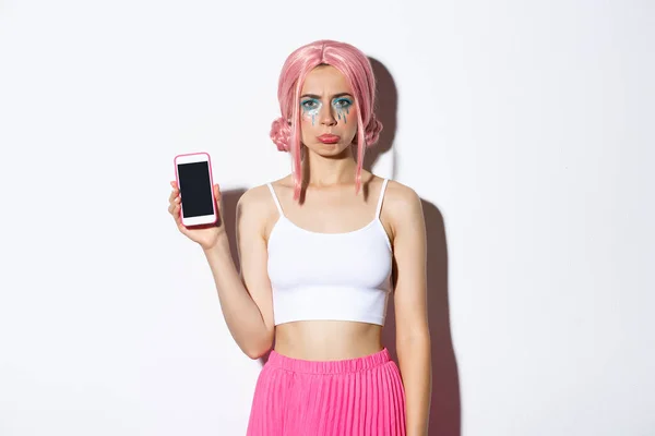 Portret dąsającej się dziewczyny narzekającej i pokazującej coś rozczarowującego na ekranie telefonu komórkowego, stojącej w różowych włosach i stroju imprezowym — Zdjęcie stockowe