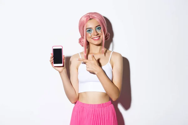 Portret pięknej modelki w różowej peruce i jasnym makijażu, wskazujący palcem na ekran telefonu komórkowego, pokazujący aplikację lub baner — Zdjęcie stockowe