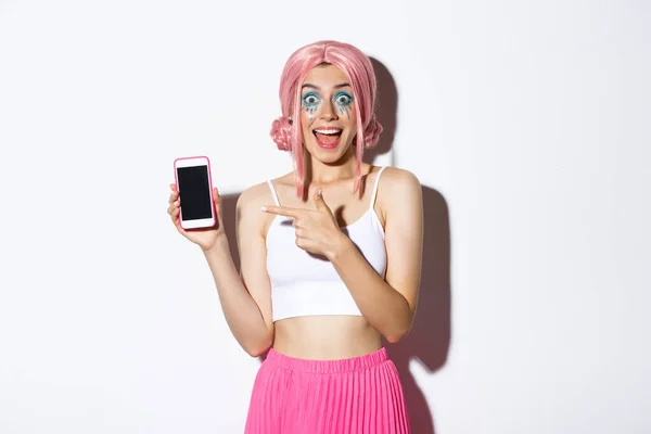 Portret podekscytowanej pięknej dziewczyny w kostiumie halloween, różowej peruce i jasnym makijażu, wskazując palcem na telefon komórkowy ze zdumioną twarzą, stojący nad białym tłem — Zdjęcie stockowe