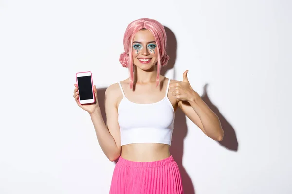 Wizerunek atrakcyjnej młodej imprezowiczki w różowej peruce, z jasnym makijażem, z kciukami do góry i ekranem telefonu komórkowego, polecam aplikację, stojącą nad białym tłem — Zdjęcie stockowe