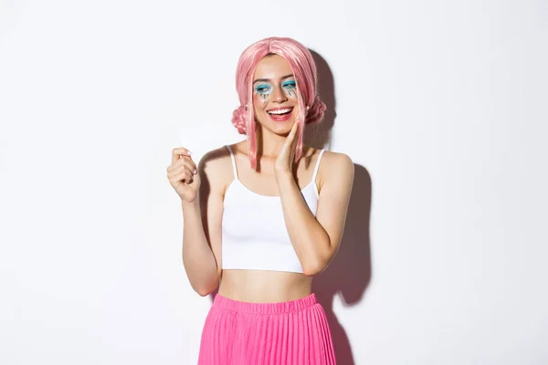 Imagem da menina elegante satisfeito em peruca rosa olhando feliz no cartão de crédito, sonhando com compras, de pé sobre fundo branco — Fotografia de Stock