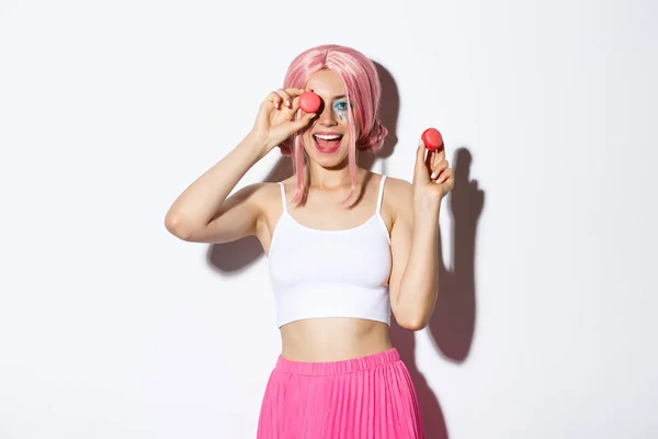 Retrato de mulher atraente alegre em peruca rosa, celebrando férias, segurando macaroons e sorrindo, de pé sobre fundo branco — Fotografia de Stock