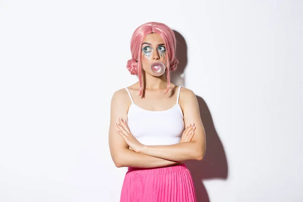 Image of confused party girl in pink wig, wigh bright makeup, blowing bubble gum and looking indecisive at upper left corner, standing over white background — Stock Photo, Image