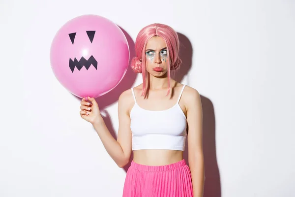 Moody schattig meisje in roze pruik, pruilen jaloers of ontstemd, staande met jack-o-lantaarn ballon, vieren Halloween, staande over witte achtergrond — Stockfoto