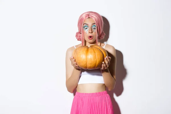 Afbeelding van aantrekkelijk meisje in roze pruik kijken verrast terwijl het houden van pompoen voor halloween partij, staande over witte achtergrond — Stockfoto