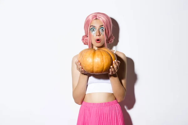 Afbeelding van aantrekkelijk meisje in roze pruik kijken verbaasd terwijl het houden van pompoen voor halloween partij, staande over witte achtergrond — Stockfoto