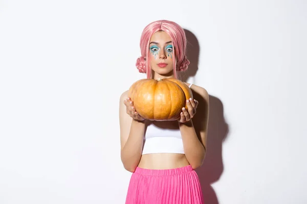 Afbeelding van mooi meisje in roze pruik en Halloween make-up, op zoek naar enthousiast op grote pompoen, staande over witte achtergrond — Stockfoto