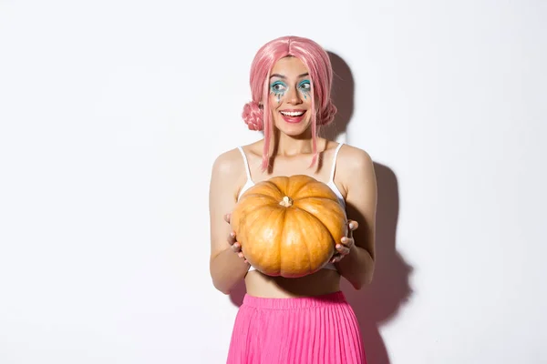 Portret van een aantrekkelijk meisje in roze pruik op zoek geamuseerd en glimlachend, het houden van pompoen voor Halloween feest, staande over witte achtergrond — Stockfoto