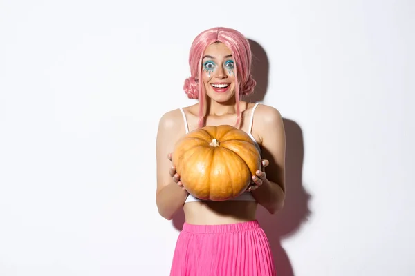 Πορτρέτο του χαρούμενος κορίτσι αναζητούν ενθουσιασμένος για το Halloween κόμμα, κρατώντας κολοκύθα, φορώντας χαριτωμένο ροζ περούκα, στέκεται πάνω από το λευκό φόντο — Φωτογραφία Αρχείου