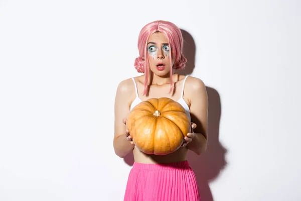 Portret van verward en bang meisje met roze pruik en heldere make-up, met pompoen voor Halloween en kijken naar links, staande over witte achtergrond — Stockfoto