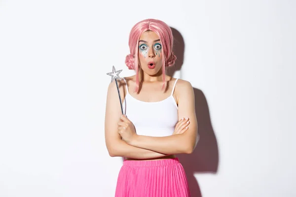 Retrato de chica sorprendida en peluca rosa, vestida de hada para la fiesta de halloween, sosteniendo varita mágica y mirando maravillado, de pie sobre fondo blanco — Foto de Stock