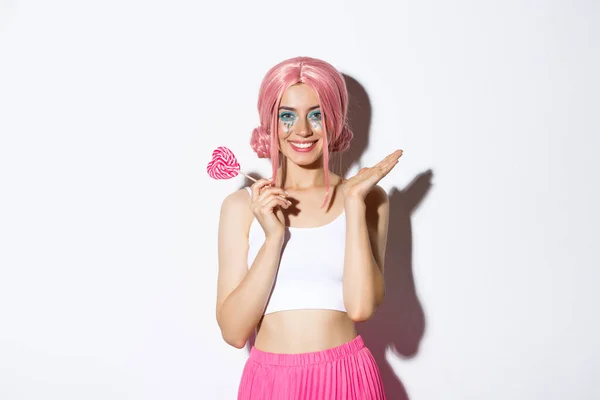 Vacker flicka ler, bär rosa peruk och älva kostym, hålla hjärtformade godis, står över vit bakgrund — Stockfoto