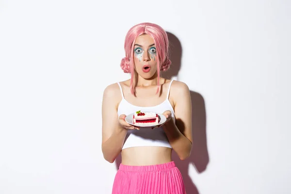 Bild von überrascht jungen Geburtstagskind in rosa Perücke, hält Kuchen auf Teller und schaut erstaunt in die Kamera, steht über weißem Hintergrund — Stockfoto