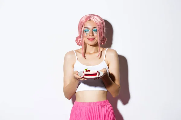 Image of beautiful girl in pink wig close eyes and smelling tasty cake, celebrating holiday, standing over white background — Stock Photo, Image