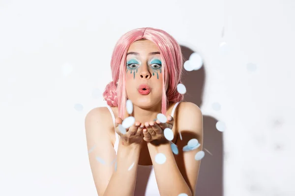 Close-up of lovely pretty party girl with pink wig and bright makeup, blowing confetti from palm, celebrating holiday, standing over white background — Stock Photo, Image