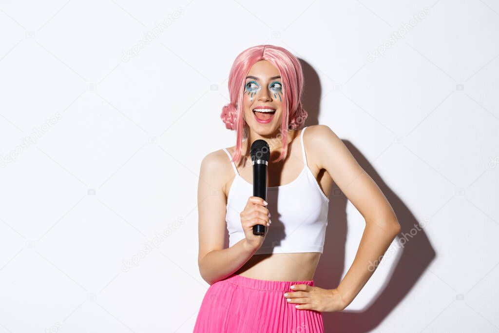 Image of beautiful smiling girl in pink wig, singing song in microphone, wearing halloween costume for party, standing over white background