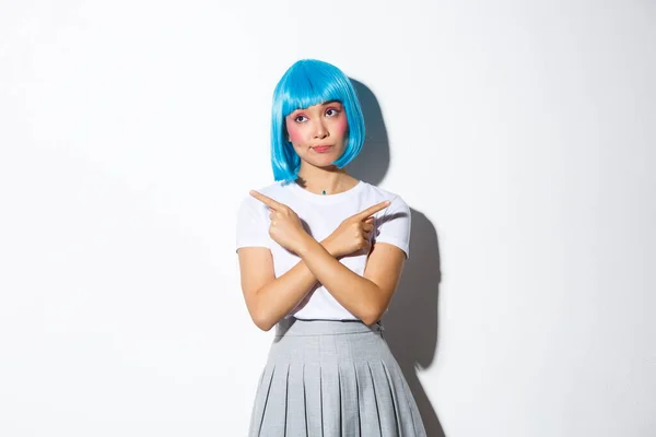 Niezdecydowana piękna azjatycka dziewczyna w niebieskiej peruce, wskazując palce bokiem i patrząc zdziwiony podczas dokonywania wyboru, ubrany na halloween, stojąc nad białym tle — Zdjęcie stockowe