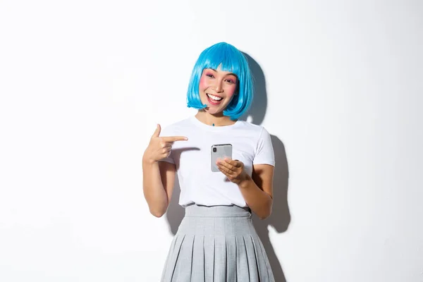 Portrait of beautiful asian girl dressed up as anime character with blue short wig, pointing finger at smartphone and smiling, standing over white background