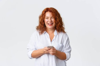 Annenin portresi, orta yaşlı kızıl saçlı kadın mesaj yazıyor, akıllı telefonla mesajlaşıyor. Kadın kameraya bakıyor, cep telefonunu tutuyor ve gülümsüyor, beyaz arka plan üzerinden uygulamayı kullanıyor.