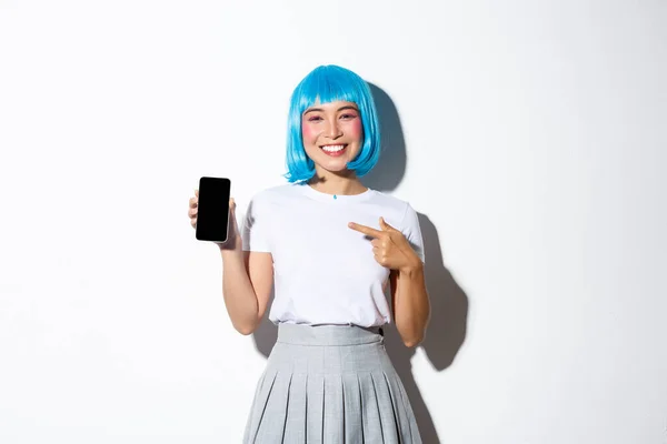 stock image Portrait of beautiful happy asian girl in blue wig pointing finger at smartphone, showing something on mobile screen, standing over white background