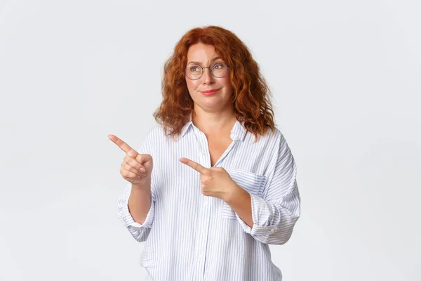 Escéptico y sin diversión, sonriente pelirroja de mediana edad mujer mirando y apuntando a algo promedio, sintiéndose reacio a la bandera promocional, no me gusta el producto, de pie fondo blanco — Foto de Stock