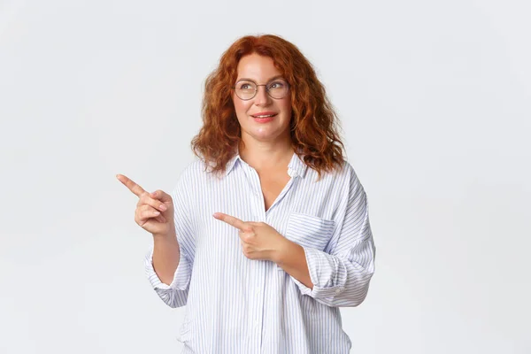 Lovely redhead middle-aged lady, female entrepreneur looking amused and pointing upper left corner. Dreamy 40s woman in glasses smiling, found excellent offer, special discount, white background — Stock Photo, Image