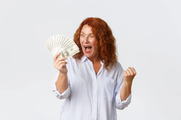 Pengar, finans och människor koncept. Triumferande, framgångsrik och vinnande rödhårig medelålders kvinna jublar över en enorm summa pengar i sina händer, dansar och skriker glatt, vit bakgrund — Stockfoto
