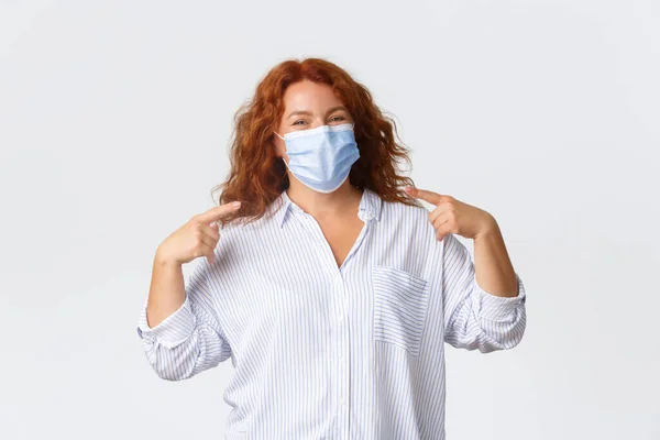 Covid-19 social distancing, coronavirus preventing measures and people concept. Cheerful smiling redhead middle-aged woman staying protected from virus, pointing at medical mask — Stock Photo, Image