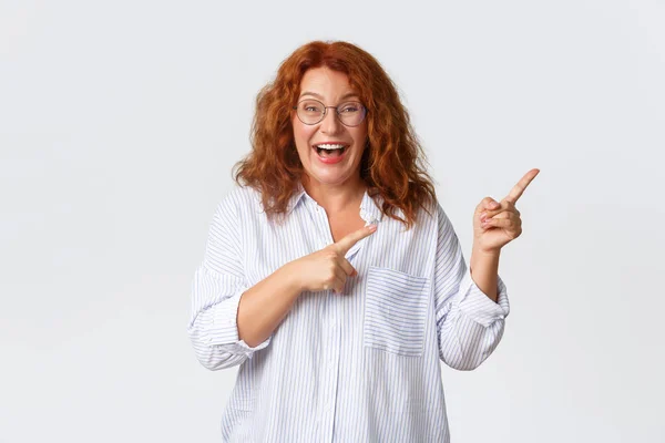 Portrait of pleasant smiling middle-aged woman with red hair, wearing glasses and blouse showing advertisement, client of company recommend product or service, pointing right — Stock Photo, Image