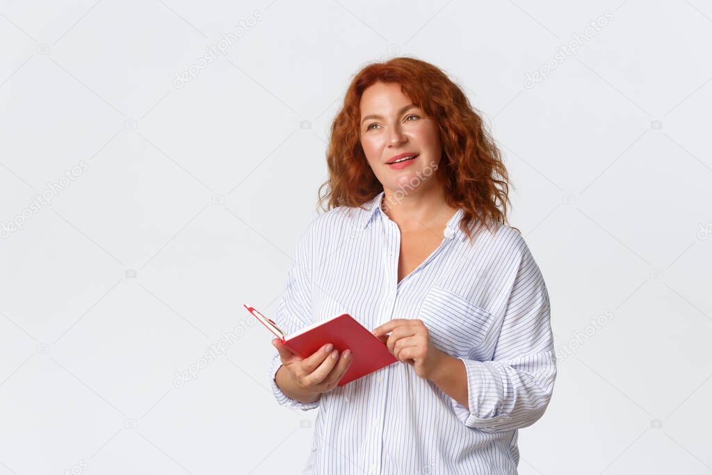 Dreamy and thoughtful middle-aged redhead woman looking away with pleased smile while holding notebook, remember something while reading diary or notes, standing white background