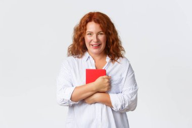 Güzel gülümseyen orta yaşlı kızıl saçlı kadın mutlu ve mutlu görünüyor, elinde neşeli bir ifadeyle kırmızı defter planlayıcısı, planlar yapıyor, notlar alıyor, çevrimiçi kurslara başlıyor, beyaz arka plan.