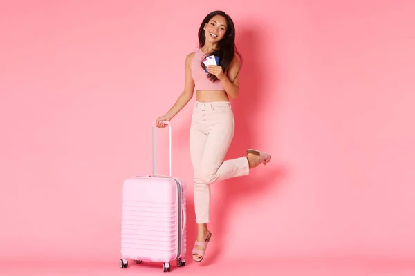 Reisen, Urlaub und Urlaubskonzept. Ganzkörperlanges fröhlich lächelndes asiatisches Mädchen, Touristin mit Flugtickets und Reisepass, vor Aufregung vor der Reise hüpfend, Koffer haltend, rosa Wand — Stockfoto