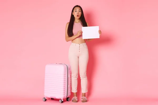 Seyahat, tatil ve tatil konsepti. Elinde kağıt parçasıyla duran eğlenceli ve büyülenmiş Asyalı güzel kız turistin pembe arka planını bavulla imzalaması. — Stok fotoğraf