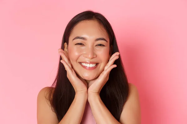 Conceito de beleza, moda e estilo de vida. Close-up de bela menina asiática tocando seu rosto e sorrindo bobo, rubor, sentindo-se atualizado, aplicar produtos de cuidados com a pele ou maquiagem, fundo rosa — Fotografia de Stock