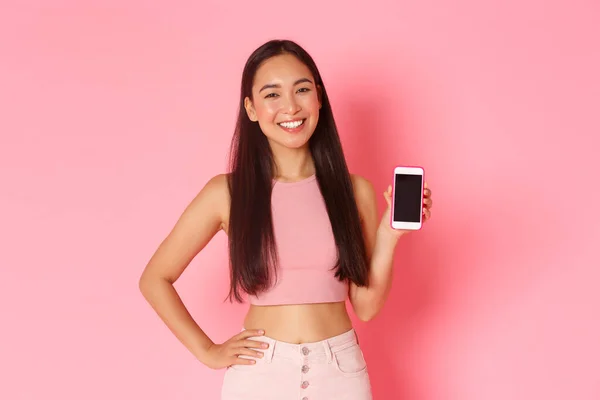 Tecnologia, comunicação e conceito de estilo de vida online. Linda menina asiática na moda em pé sobre fundo rosa com telefone celular, mostrando aplicativo ou tela do smartphone banner, fundo rosa — Fotografia de Stock