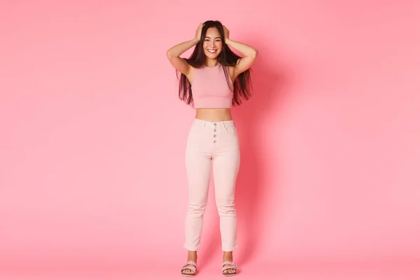 Moda, beleza e estilo de vida conceito. Comprimento total retrato de atraente asiático menina no glamour roupa tocando cabelo e sorrindo amplamente, alegrando-se com novo incrível corte de cabelo, fundo rosa — Fotografia de Stock