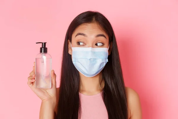 Covid-19 salgını, koronavirüs ve sosyal uzaklık kavramı. Tıbbi maske takmış şirin ve aptal Asyalı bir kız el dezenfektanına bakıyor, antiseptik kullanıyor, pembe arka plan kullanıyor. — Stok fotoğraf